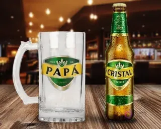 Jarra de cerveza de vidrio con diseño sublimado 'Para un Papá Increíble' en tonos dorados y verdes, al estilo de Cerveza Cristal, al lado de una botella de Cerveza Cristal con gotas de condensación.