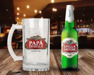 Shopper cervecero transparente con diseño 'Papá Estrella' en rojo y blanco, inspirado en Stella Artois, junto a una botella de cerveza Stella Artois fría sobre una superficie de madera rústica.