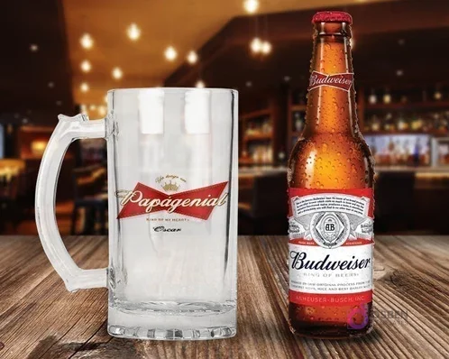 Shopper de vidrio personalizado con el diseño 'Papagenial King of my heart' en rojo, blanco y dorado, junto a una botella de Cerveza Budweiser con condensación en un ambiente de bar.