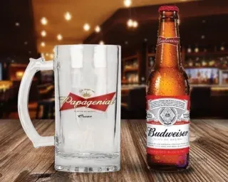 Shopper de vidrio personalizado con el diseño 'Papagenial King of my heart' en rojo, blanco y dorado, junto a una botella de Cerveza Budweiser con condensación en un ambiente de bar.