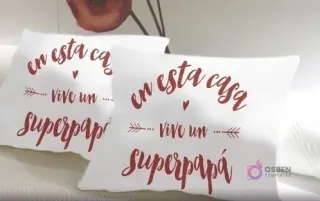 Dos cojines blancos con la frase 'En esta casa vive un superpapá' en color vino tinto, adornada con flechas y pequeños corazones, en un estilo manuscrito y emotivo.