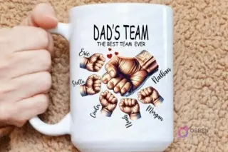 Mano sosteniendo una taza grande de sublimación con el diseño 'Dad's Team The Best Team Ever' y siete puños personalizados con nombres, resaltando la capacidad de añadir más miembros de la familia.