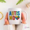 Manos sujetando una taza blanca con la palabra 'AUTISMO' en colores vivos y un corazón con piezas de puzzle. Rodeada de hilos y cuentas doradas.