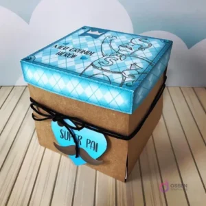 Caja explosiva cerrada en color kraft con tapa decorada en tonos azules y blancos, atada con lazo negro y etiqueta 'Super Pai', diseño para el Día del Padre.