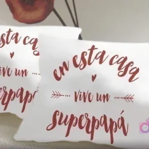 Dos cojines blancos con la frase 'En esta casa vive un superpapá' en color vino tinto, adornada con flechas y pequeños corazones, en un estilo manuscrito y emotivo.