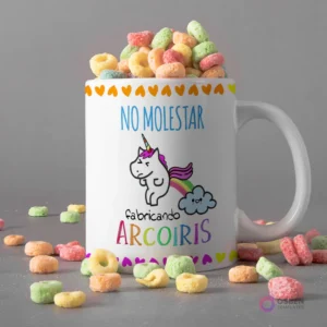Taza blanca con diseño de unicornio haciendo caca de arcoíris y frase 'NO MOLESTAR fabricando ARCOIRIS', rodeada de cereal Fruity Loops de colores.