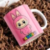 Taza de cerámica rosa con diseño del personaje Labubu de traje peludo rosa, con el nombre Labubu en letras coloridas, sobre papel picado marrón.