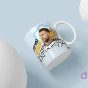 Mockup de taza blanca para sublimación con diseño de Lionel Messi y la frase '¿Qué Mirás Bobo? Andá Pa' Allá'. El diseño incluye detalles de la bandera argentina y el sol, sobre un fondo azul claro con dos esferas blancas. La taza está flotando en un ángulo dinámico.