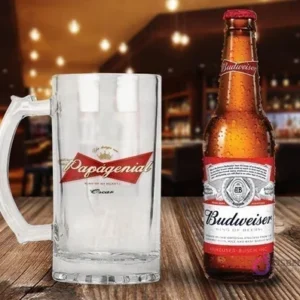Shopper de vidrio personalizado con el diseño 'Papagenial King of my heart' en rojo, blanco y dorado, junto a una botella de Cerveza Budweiser con condensación en un ambiente de bar.
