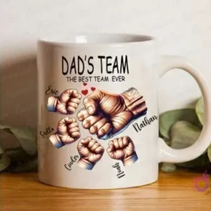 Taza blanca de sublimación con diseño 'Dad's Team The Best Team Ever' featuring cinco puños personalizables con nombres 'Eric, Stella, Cinder, Nathan, Noah' sobre un fondo desenfocado.